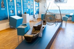 Fragment wystawy z planszami i małymi gablotami, na środku zabytkowa drewniana łódka z wiosłem, nad nią zawieszone sieci