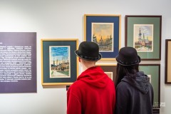 Dwie osoby w czarnych kapeluszach typu melonik na głowie patrzą na obrazy wiszące na wystawie