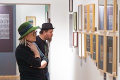 Kobieta w zzielonym kapeluszu i mężczyzna w czarnym kapeluszu typu melonik oglądają obrazy na wystawie