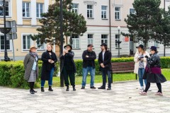 Na zwenątrz stoi siedem osób. W tle zieleń, a dalej fragment budynku ostrołęckiego ratusza