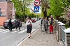Kilka osób idzie po chodniku w starej części miasta.  Z prawej strony ogrodzenie, z lewej fragment budynku i osoby przechodzące przez jednię, w tle zabudowania i zieleń