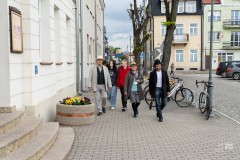 Kilka osób idzie po chodniku w starej części miasta. Z lewej strony elewacja budynku, z prawej brukowa ulica
