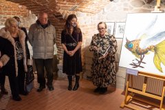Dyrekcja Muzeum, autorka wystawy oraz uczestnicy wernisażu stoją podczas otwarcia wystawy w muzealnych piwnicach. Z prawej strony na sztaludze fragment jednej z grafik.