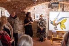 Dyrekcja Muzeum, autorka wystawy oraz uczestnicy wernisażu stoją podczas otwarcia wystawy w muzealnych piwnicach. Z prawej strony na sztaludze fragment jednej z grafik.