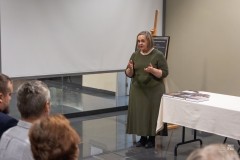 Kobieta stojąc przemawia do widowni, z której widocznych jest kilka osób