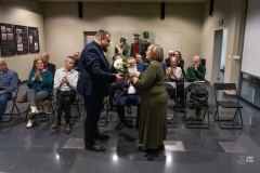 Kobieta i mężczyzna stoją naprzeciw siebie, następuje wręczenie kwiatów. W tle widownia w liczbie kilkunastu osób. Z lewej strony na ścianie fragment wystawy planszowej.