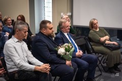 W sali  na widowni siedzi kilka osób. Mężczyzna w pierwszym rzędzie trzyma bukiet kwiatów.