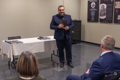 Mężczyzna stojąc trzyma książkę i przemawia do zgromadzomnych na widowni. Widoczne dwie osoby. Z prawej strony, na ścianie, fragment wystawy planszowej.