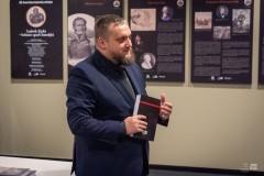 Mężczyzna stoi trzymając w dłoniach książkę. W tle na ścianie fragment wystawy planszowej
