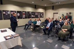 Mężczyzna stojąc przemawia do widowni, na której widać kilkanaście siedzących osób. W tle, na ścianie, wystawa planszowa