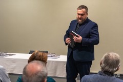 Mężczyzna stojąc trzyma książkę i przemawia do zgromadzomnych na widowni. Widoczne kilka osób