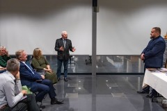 Dwóch mężczyzn stoi, jeden przemawia, z lewej strony fragment widowni, siedzi kilka osób