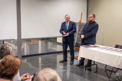 Dwóch mężczyzn stoi z boku stołu, przez widownią. Widoczny niewielki fragment widowni