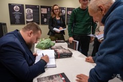 Autor książki siedzi przy stole i podpisuje egzemplarze. W kolejce czeka kilka osób.