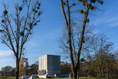 Dwa wysokie drzewa, pomiędzy drzewami, w oddali Pomnik Mauzoleum oraz trzy flagi na masztach