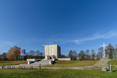 Pomnik Mauzoleum w oddali. Po prawej stronie wysoki krzyż, po lewej trzy maszty z flagami Mazowsza, Polski i Unii Europejskiej
