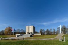 Pomnik Mauzoleum w oddali. Po prawej stronie wysoki krzyż, po lewej trzy maszty z flagami Mazowsza, Polski i Unii Europejskiej