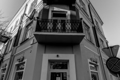 Zdjęcie czarno-białe. Róg budynku, na środku fragment drzwi, na piętrze balkon