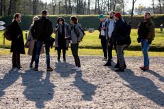 Dziewięć osób, kobiety i mężczyźni. Niektórzy trzymają w dłoniach aparaty fotograficzne. W oddali zielony teren, wznosi się wał, fragmenty drzew