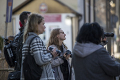 Kilka osób patrzy w prawą stronę, niektórzy trzymają w dłoniach aparaty fotograficzne. W tle zabudowania.