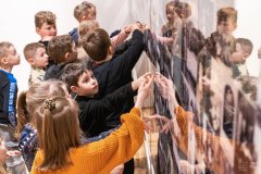 Dzieci stoją przy ścianie, na której eksponowane są zabytkowe fotografie. Wiele z dzieci wskazuje dłonią na poszczególne zdjęcia.
