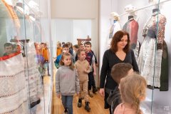 Pracownica Muzeum prowadzi grupę dzieci między dużymi gablotami, w których prezentowane są stroje kurpiowskie