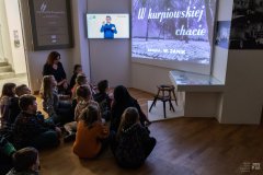 W zaciemnionym pomieszczeniu dzieci oraz pracownica Muzeum siedzą na podłodze i oglądają projekcję czarnobiałego filmu. Na planszy tytułowej filmu napis: „W kurpiowskiej chacie”. W tle monitor na którym mężczyzna  komunikuje się w języku migowym.