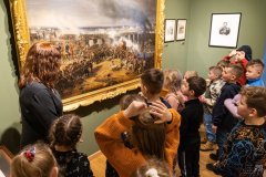 Dzieci stojąc patrzą na wielki obraz w zdobionej ramie. Z dziećmi jest pracownica Muzeum.