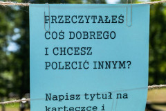 Na poziomych sznurkach przymocowana kartka z napisem „Przeczytałeś coś dobrego i chcesz polecić innym? Napisz tytuł na karteczce i przyklej do sznurka”. W tle zieleń, drzewa.