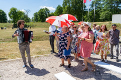 Akordeonista grając prowadzi śpiewający podczas spaceru chór. Niektóre osoby z chóru trzymają chroniące przed słońcem biało-czerwone parasole. W tle zieleń, drzewa.