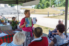 Kobieta trzyma w dłoniach książki, prezentując je przed widownią. W tle teren ostrołęckich Fortów Bema.