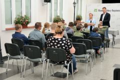 kilkanaście osób w tym harcerze siedzą na krzesełkach i słuchają przemowy prowadzących.