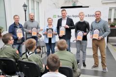 sześć osób stoi z nagrodami i pozuję do zdjęcia, przed nimi siedzą harcerze.