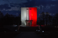 duża budowla nocą, pomnik mauzoleum poświęcony bohaterom poległym 26 maja 1831 roku , podświetlony polskimi barwami narodowymi  w kolorze biało-czerwonym, w tle kilkanaście drzew , na środku pomnika wejście do krypty, nad wejściem duży krzyż
