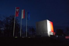 ogromna betonowa budowla nocą podświetlona barwami narodowymi biało-czerwonymi, pomnik mauzoleum poświęcony  bitwie pod Ostrołęką 26 maja 1831 roku, przed pomnikiem powiewają trzy maszty z chorągwiami, flaga województwa mazowieckiego w kolorze czerwonym z umieszczonym po lewej wizerunkiem orła srebrnego o dziobie i szponach, następna biało-czerwona flaga Polski i flaga europejska przedstawiająca okrąg złożony z dwunastu złotych gwiazd pięcioramiennych na lazurowym tle  