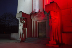 fragment betonowej budowli nocą z podświetleniem  barw narodowych biało-czerwonych, pomnik bohaterom poległym 26 maja 1831 roku, po lewej i prawej stronie wejścia do krypty stoją rzeźby żołnierzy, w tle widoczne drzewa  