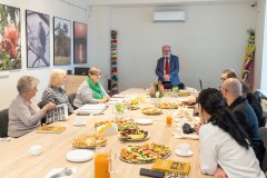 Po obu stronach dużego stołu obradują uczestnicy  zebrania, jeden obradujący stoi za stołem na końcu, za nim po lewej i prawej stronie w rogu sali stoją wielkie kolorowe palmy wielkanocne. W tle na ścianie po lewej stronie  wiszą ilustracje zwierząt i przyrody.  
