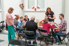 Uczestnicy warsztatów, dorośli i dzieci, siedzą przy stole i wykonują maki z bibuły