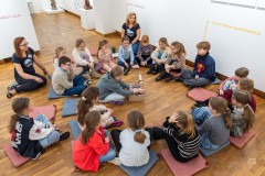 W sali wystawowej kilkanaścioro dzieci siedzi na podłodze tworząc duże koło. Przy dzieciach siedzą dwie muzealniczki. Jedno dziecko siedzące w środku koła zagląda do pudełka z wieczkiem