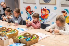 Cztery dziewczynki siedzą przy dużym stole i wykonują wycinanki. Piąta dziewczynka stoi po lewej stronie przy stole i spogląda na pozostałe.