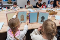 Dwie dziewczynki siedzące przy stole oglądają swoje albumy z wycinankami. W tle inne dzieci siedzące przy stole i wykonujące wycinanki.