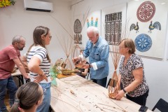 kilka osób robi plecionki, w tle obrazy z wycinankami kurpiowskimi