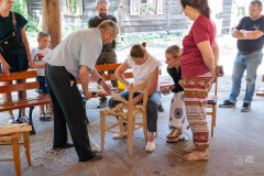 kobieta robi plecionkę podczas gdy inni się na to spoglądają w tle stara wiejska chatka
