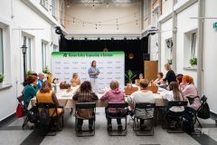 osoba na scenie przemawia do kilku innych osób w tle ściana z napisem i logotypem MKK oraz logotypem Mazowsze