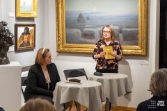 Dwie kobiety prowadzące spotkanie. Jedna z kobiet stojąc trzyma w dłoniach książkę i przemawia do zebranych w sali wystawowej uczestników spotkania, druga siedzi przy okrągłym stoliku. W tle, na ścianie, wiszą obrazy.