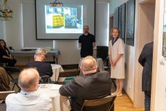Kilkunastu uczestników słucha przemówienia otwierającego spotkanie promocyjne książkę "Macvra" przez panią stojącą po prawej stronie sali, tuż za nią stoi mężczyzna za którym na ekranie widnieje promocyjna książka.