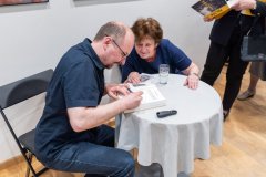 Siedząc przy stoliku autor podpisuje książkę siedzącej obok kobiecie. 