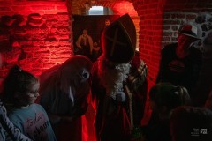 W piwnicy dzieci, duch, św. Mikołaj i kobieta w kapeluszu. Pomieszczenie oświetlone czerwonym światłem