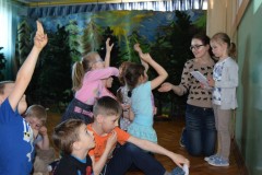 Dzieci podnoszą ręce, przed nimi stoi dziewczynka oraz pani.