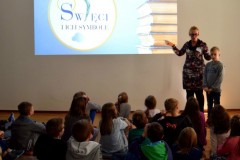 Kobieta wskazuje na wyświetlacz, dzieci oglądają siedząc.
